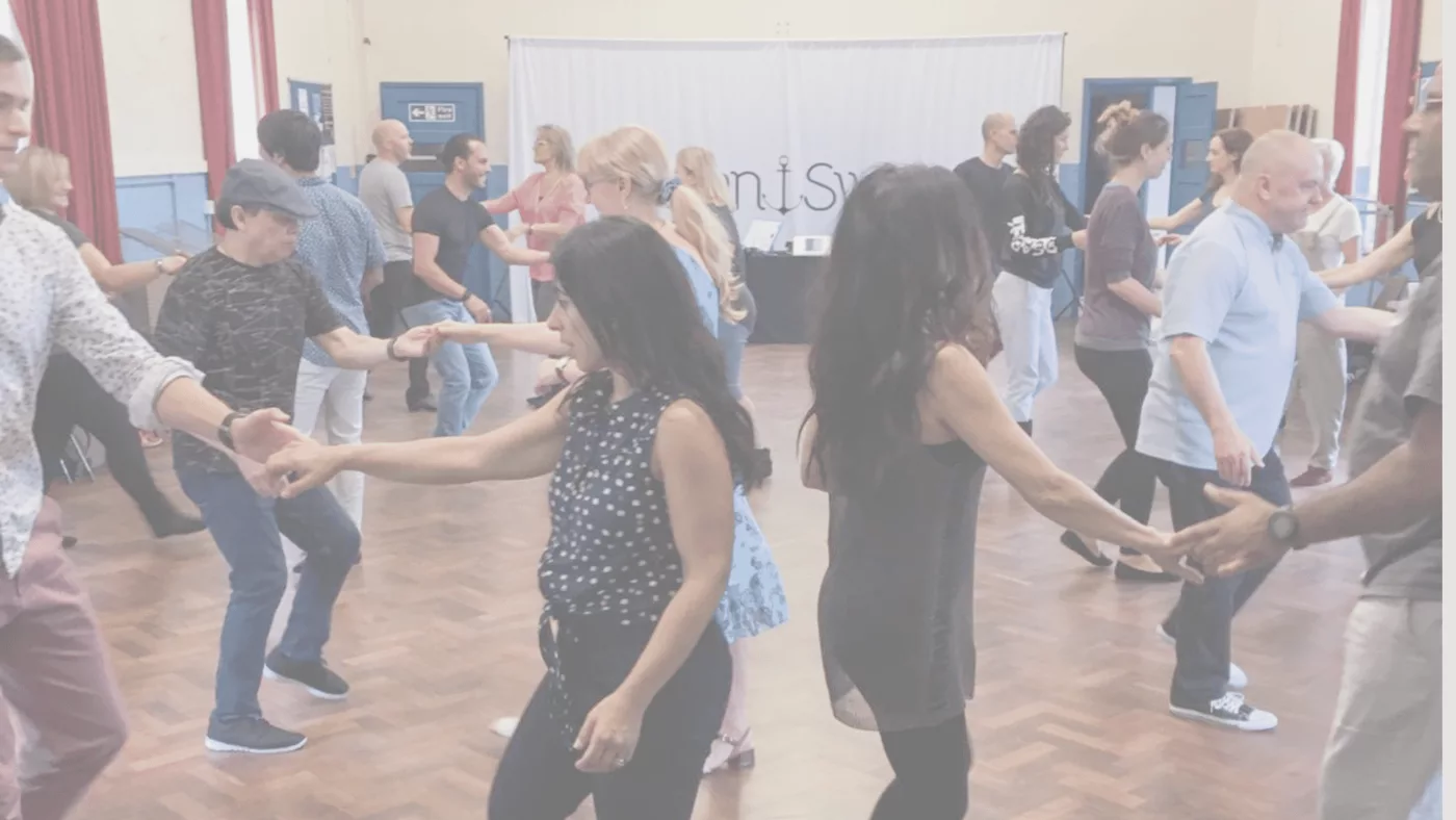 A west coast swing group of men and women dancing west coast swing dancers in a church hall with a dark parquet floor.